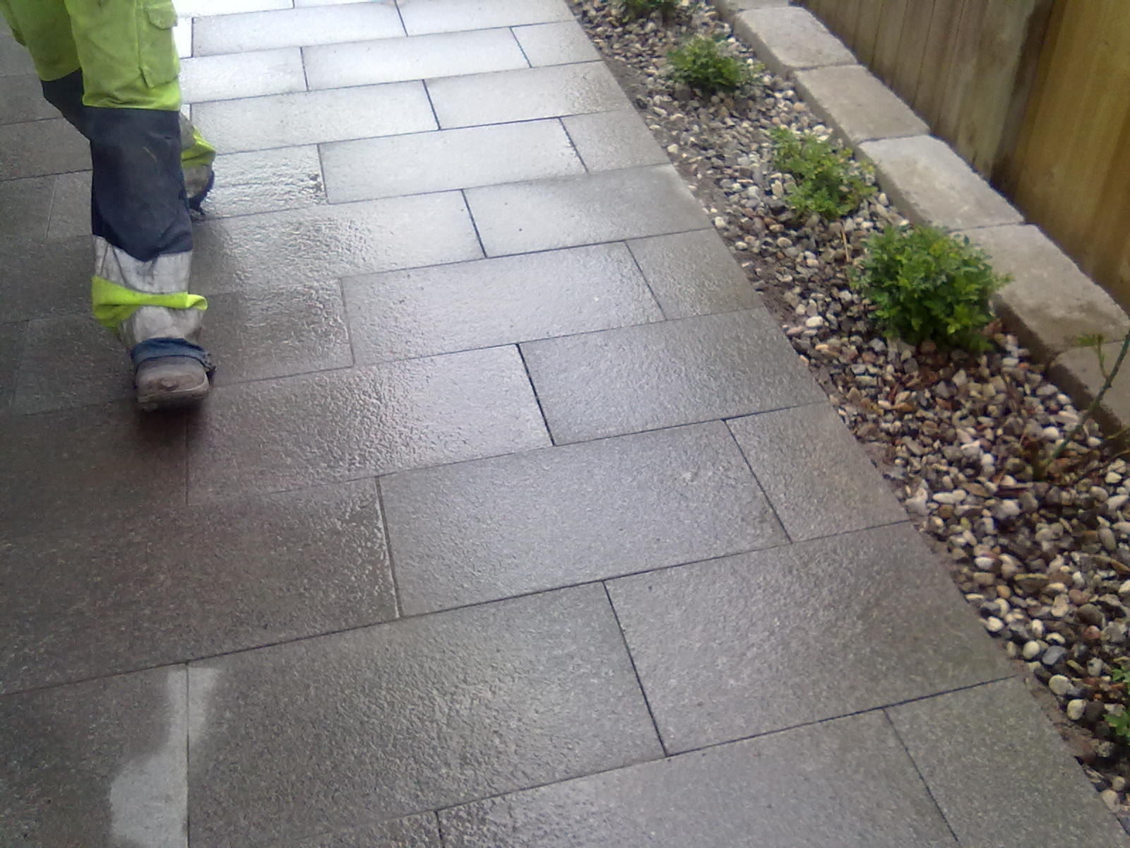 Świeżo ułożona, mokra nawierzchnia z szarych płyt granitowych, widoczne fragmenty stroju roboczego osoby wykonującej prace i obrzeże z kamyków oraz roślin.
