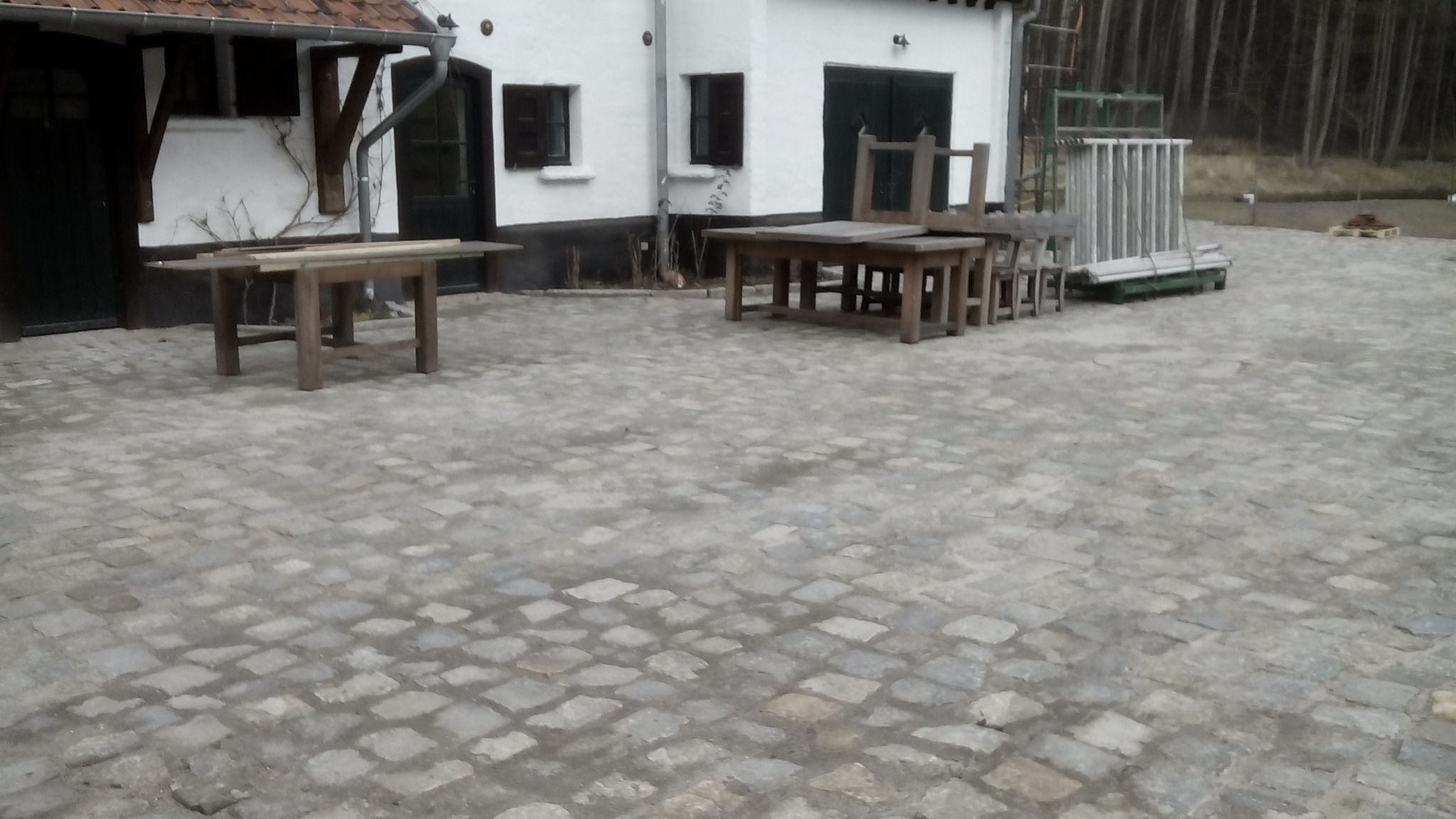 Szara kostka granitowa na podwórku z białym budynkiem w tle, widoczne drewniane stoły i krzesła oraz rusztowanie.