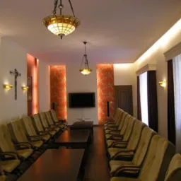 Wnętrze sali konferencyjnej z rzędami krzeseł, stołami, oświetleniem sufitowym w stylu Tiffany oraz dekoracyjną tapetą i krzyżem na ścianie.