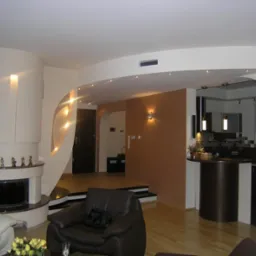 Nowoczesne wnętrze salonu z kominkiem i otwartą kuchnią, widoczne gładzie na ścianach, oświetlenie punktowe i drewniana podłoga.