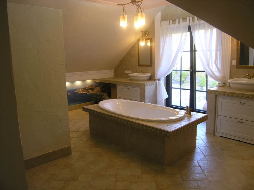 Jasna łazienka na poddaszu ze skośnym sufitem, wolnostojąca wanna na podeście z płytek, dwie umywalki nablatowe z szafkami, okno z białymi zasłonami i fototapeta z motywem morskim.