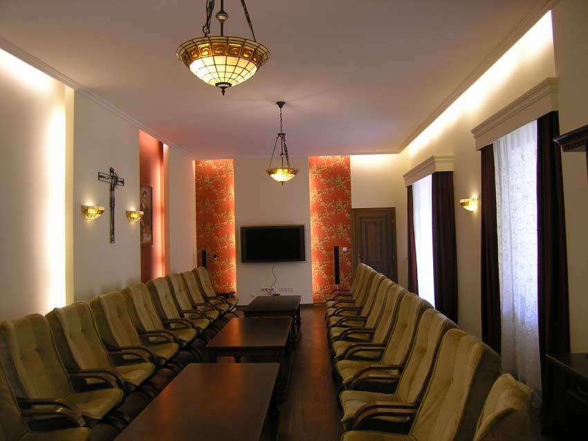 Wnętrze sali konferencyjnej z rzędami krzeseł, stołami, oświetleniem sufitowym w stylu Tiffany oraz dekoracyjną tapetą i krzyżem na ścianie.