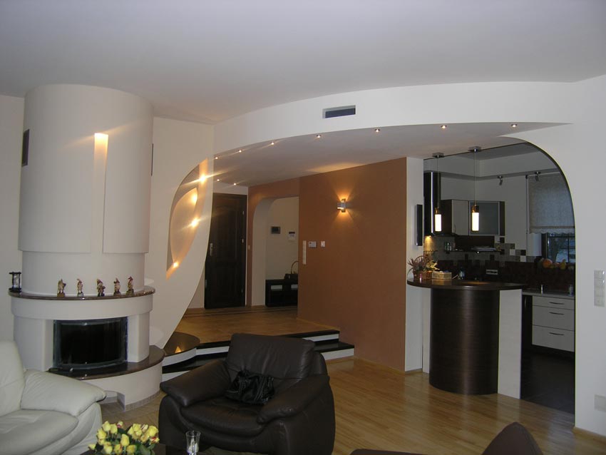 Nowoczesne wnętrze salonu z kominkiem i otwartą kuchnią, widoczne gładzie na ścianach, oświetlenie punktowe i drewniana podłoga.