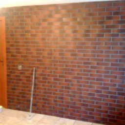 Ściana wykończona płytami imitującymi czerwoną cegłę, widoczny fragment drzwi i podłogi z płytek, w tle kontakt elektryczny i pionowa rurka