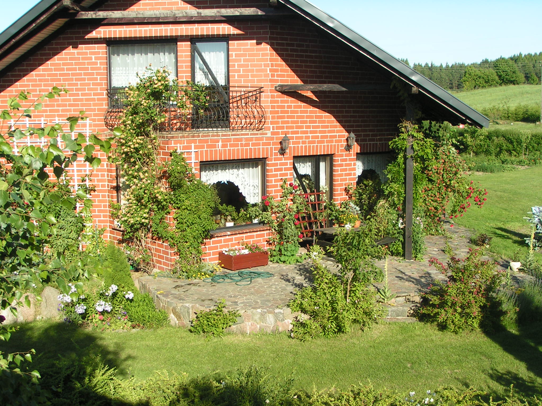 Ceglany dom z balkonem i bujną roślinnością pnącą się po ścianach, otoczony zielonym ogrodem z kamienną ścieżką i rabatami kwiatowymi.