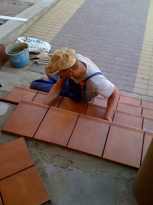 Pracownik w ubraniu roboczym układa terakotowe płytki schodowe na zaprawie cementowej, widoczny fragment chodnika z kostki brukowej, wiadro i worek z zaprawą w tle.
