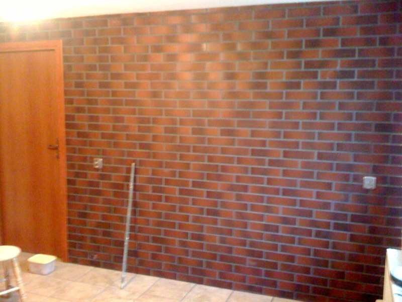 Ściana wykończona płytami imitującymi czerwoną cegłę, widoczny fragment drzwi i podłogi z płytek, w tle kontakt elektryczny i pionowa rurka