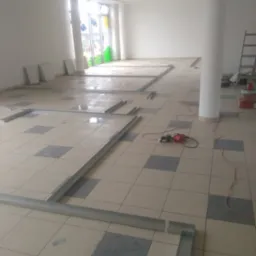 Montaż stelażu pod płytę karton gips na podłodze w jasnym pomieszczeniu z kolumną, widoczne profile, wiertarka i płytki podłogowe.