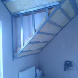 Skośne poddasze w trakcie ocieplania wełną mineralną, widoczna metalowa konstrukcja nośna, grzejnik, wiadro z zaprawą i mieszadło elektryczne w tle.