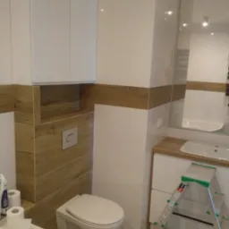 Nowoczesna łazienka z białymi błyszczącymi płytkami, drewnopodobnymi akcentami, podwieszaną toaletą i drabinką Dexter opartą o szafkę umywalkową.