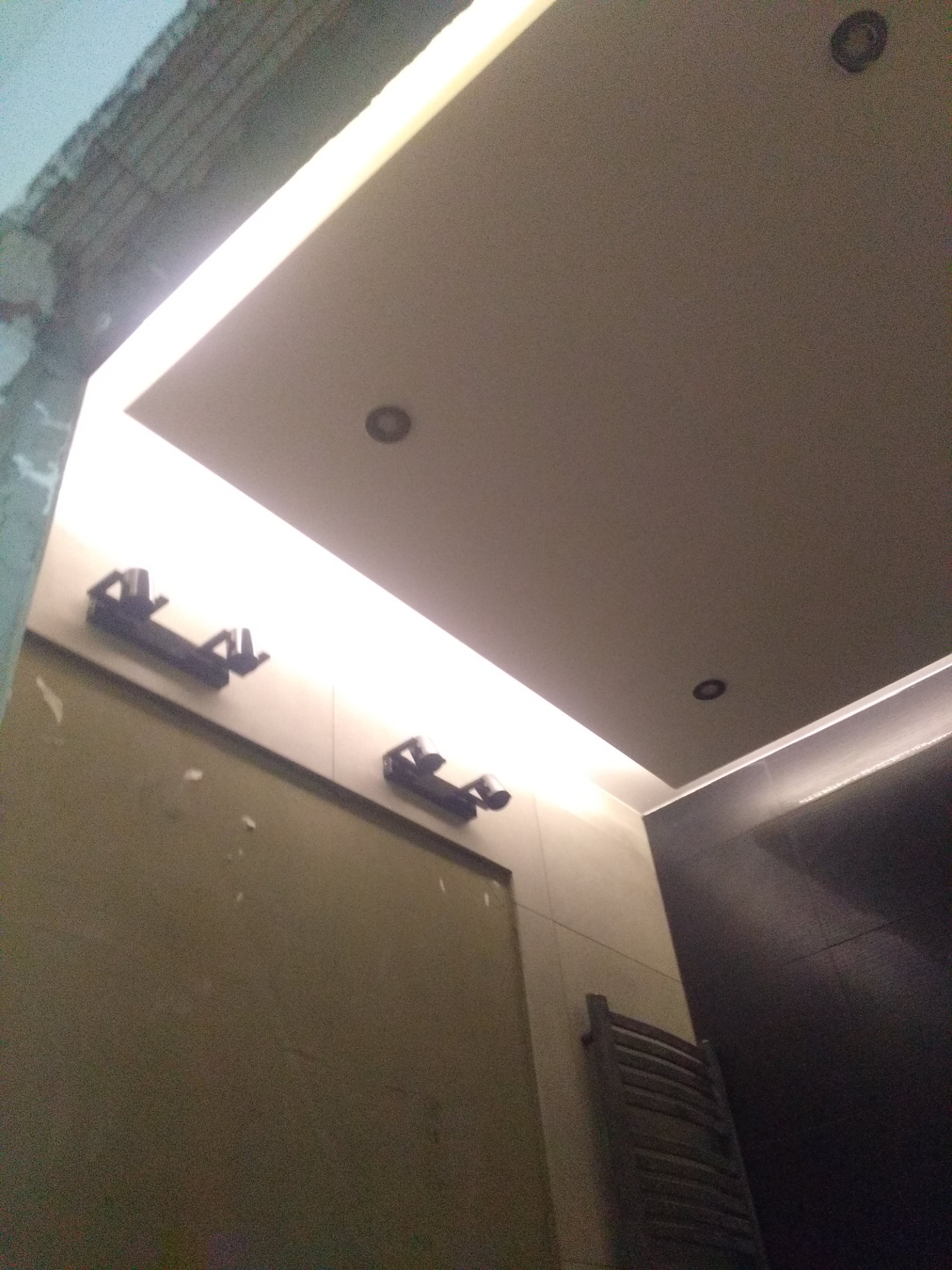 Niedokończona łazienka z widoczną instalacją oświetlenia LED w suficie podwieszanym, fragment surowego muru na górze po lewej i grzejnik drabinkowy na ścianie.