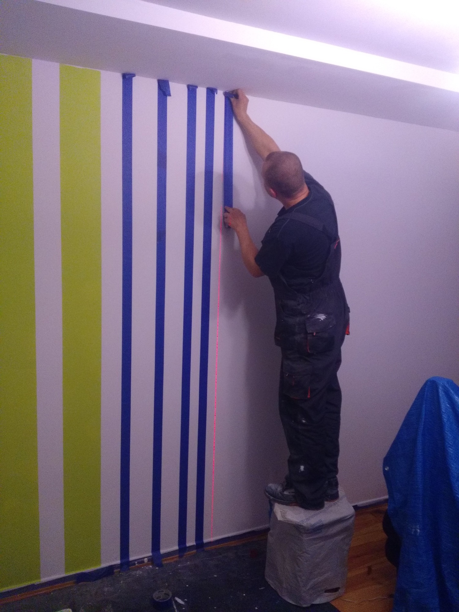 Malowanie ścian w pionowe pasy: pracownik na podwyższeniu przykleja taśmę malarską, widoczna zielona i biała pasma oraz laserowy wskaźnik pionu.