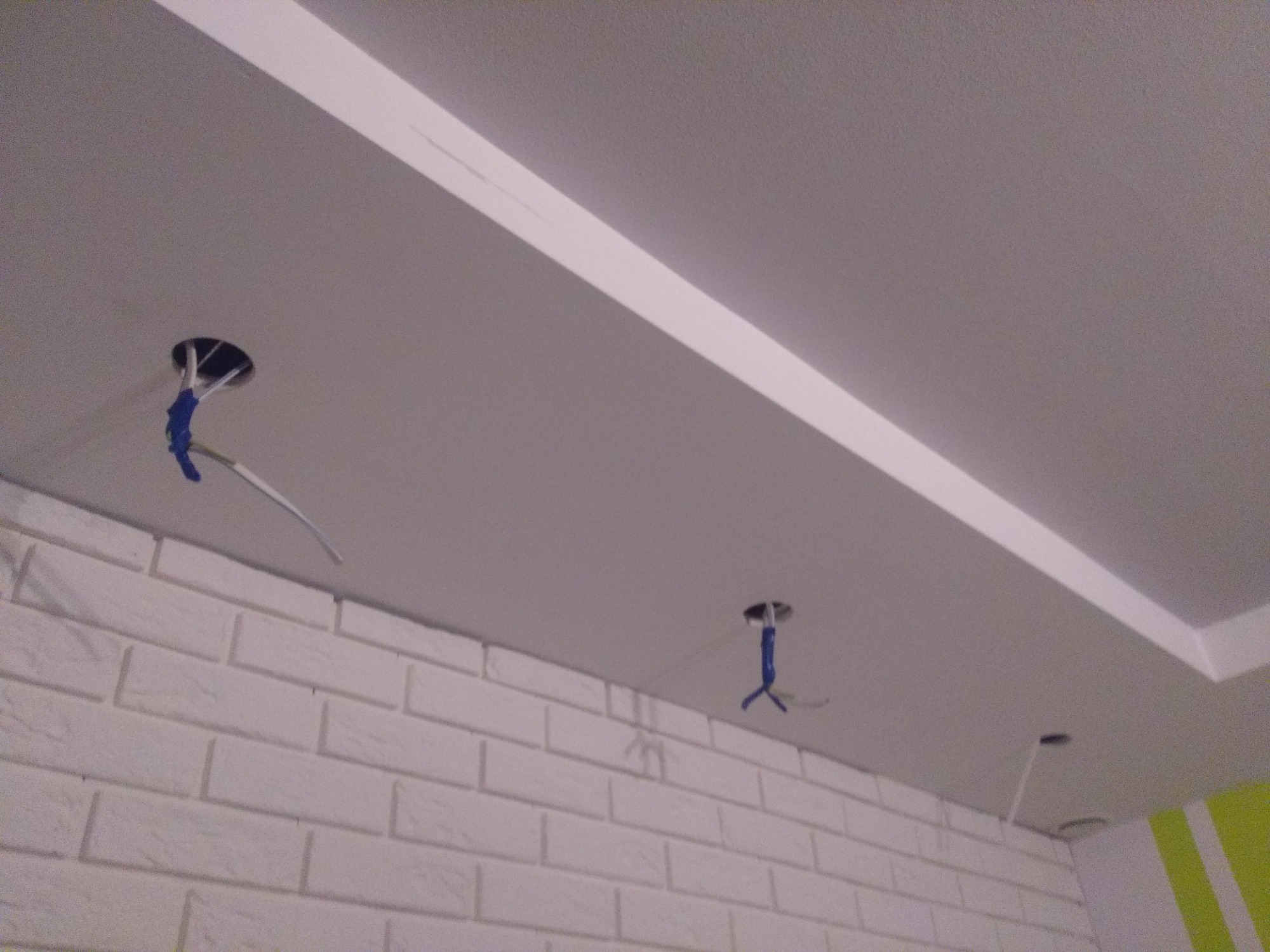 Biały sufit z zamontowanym oświetleniem LED w formie wpuszczonej listwy, widoczne przewody elektryczne wystające z otworów pod przyszłe punkty świetlne, ściana wykończona białą cegłą dekoracyjną.