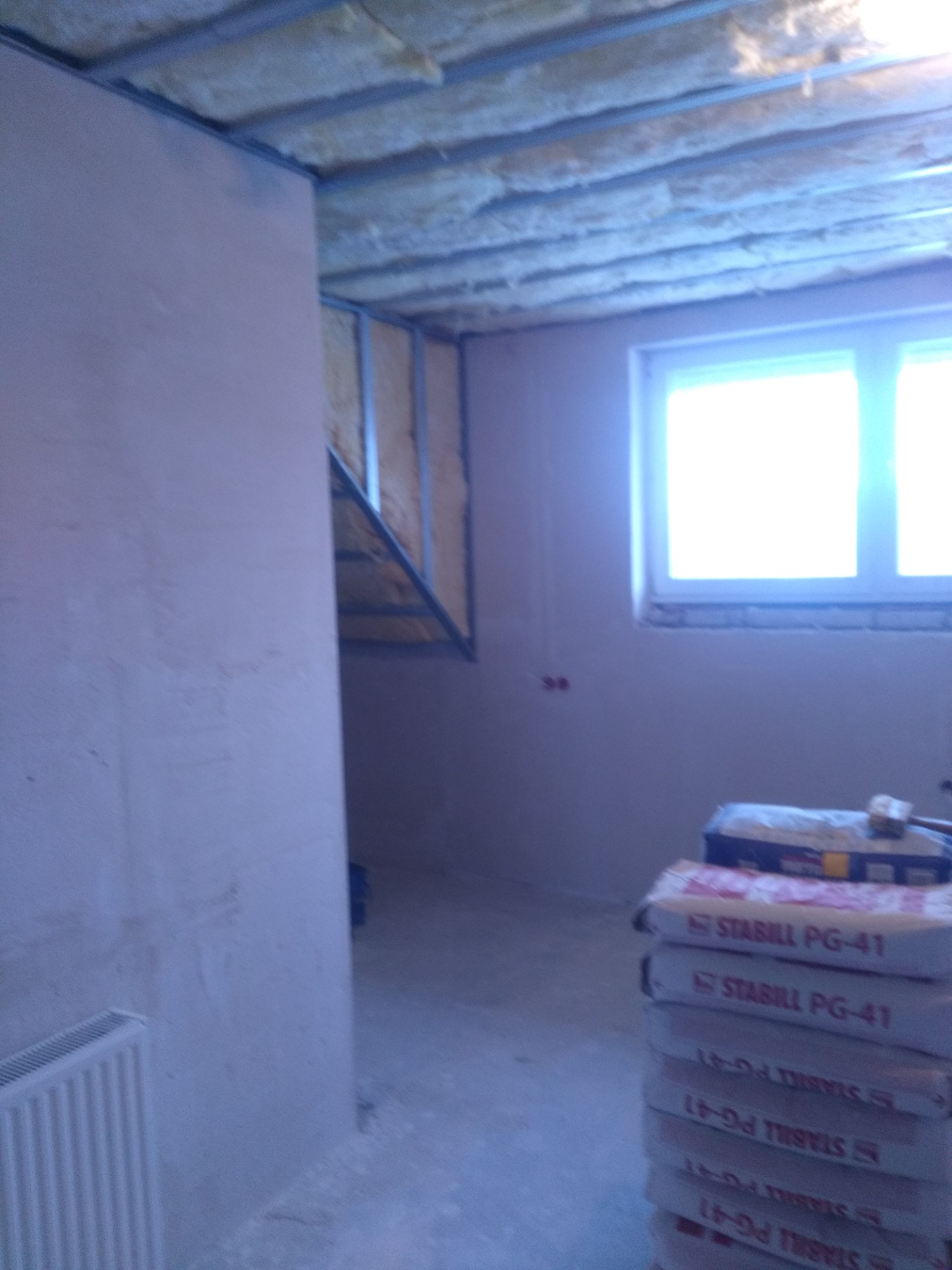 Poddasze w trakcie ocieplania, widoczne wełna mineralna między krokwiami, stelaż pod płytę karton-gips, worki z gładzią szpachlową Stabili PG-41 i grzejnik w surowym stanie.