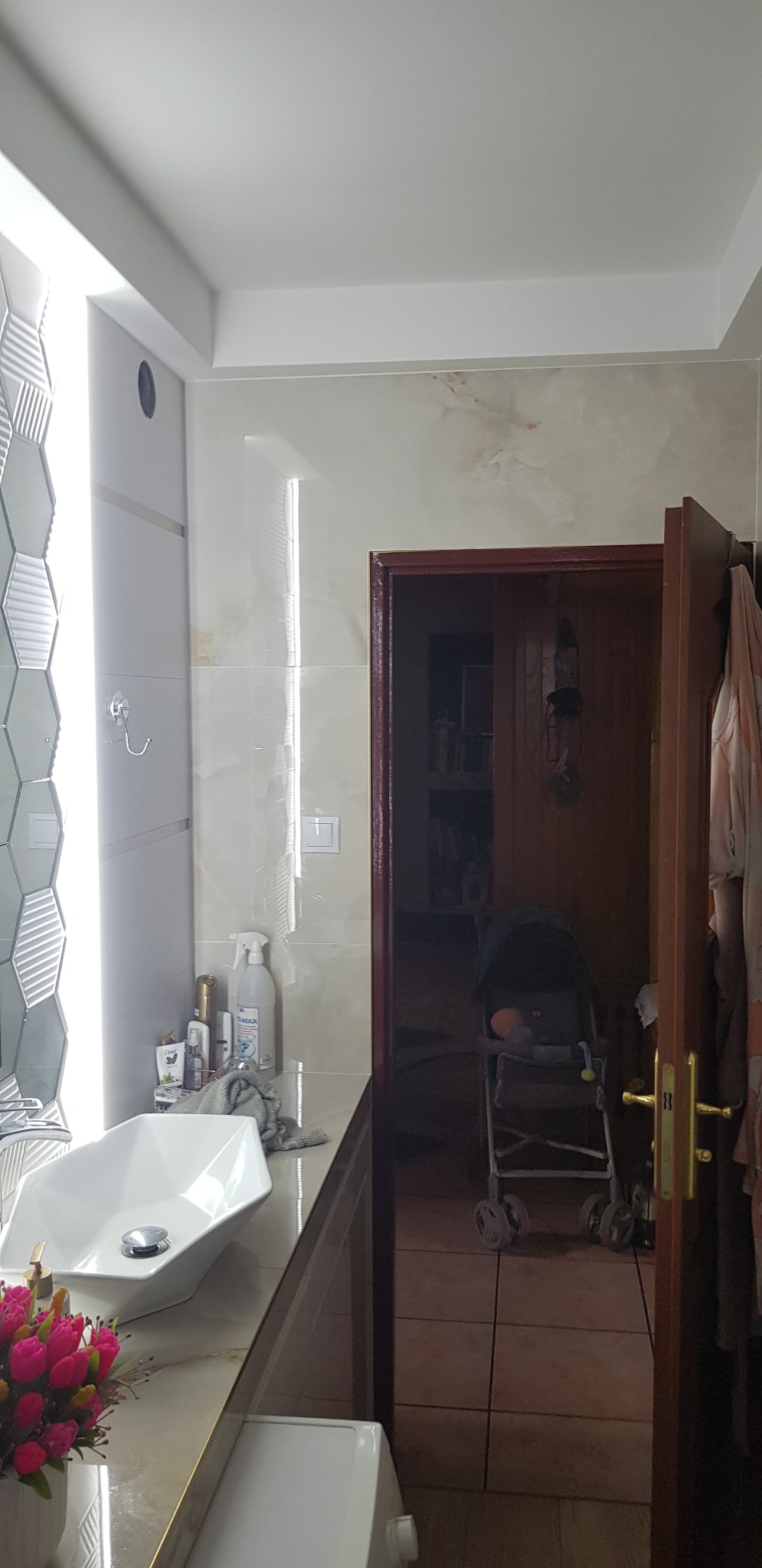 Nowoczesna łazienka z umywalką nablatową, heksagonalnymi płytkami na ścianie i widokiem na pokój z wózkiem dziecięcym w tle.