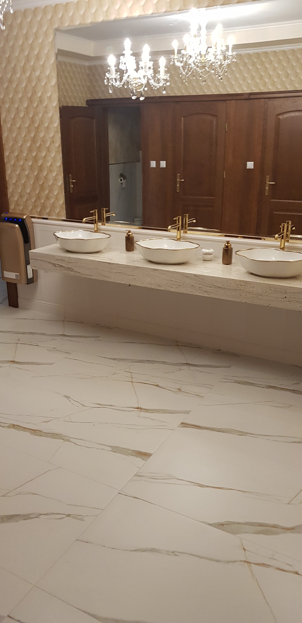 Elegancka toaleta publiczna z trzema umywalkami w stylu glamour, złotymi bateriami i marmurową posadzką w odcieniach bieli i beżu, oświetlona kryształowym żyrandolem.