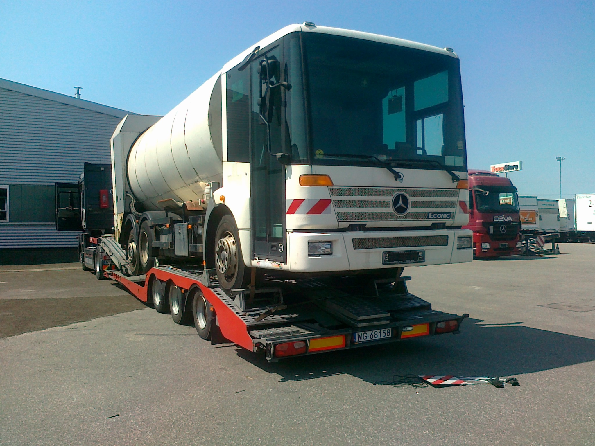 Ciężarówka-cysterna Mercedes Econic na czerwonej platformie niskopodwoziowej, widok z przodu, tablica rejestracyjna WG 68158, budynek magazynowy w tle.