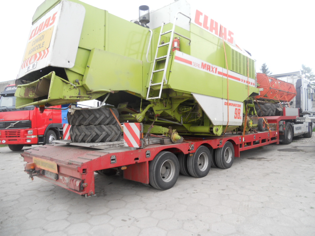 Zielono-biały kombajn zbożowy Claas Dominator 98S zabezpieczony pasami na czerwonej platformie niskopodwoziowej z dodatkowymi oponami, widoczny czerwony ciągnik siodłowy Volvo z przodu i biały...