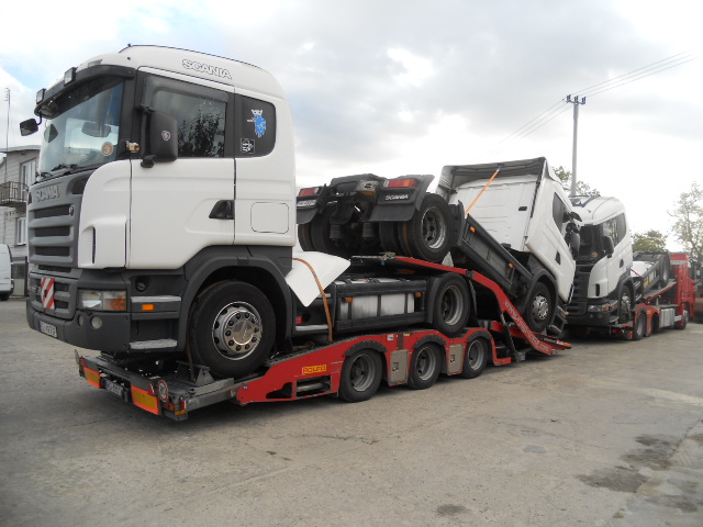 Czerwona laweta z trzema białymi ciągnikami siodłowymi marki Scania, ułożonymi jeden na drugim, przewożonymi na platformie z wieloma osiami.