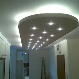 Sufit podwieszany z oświetleniem punktowym w kształcie półkola, listwy dekoracyjne przy ścianach, wnętrze w trakcie wykańczania, widoczna drabina oparta o ścianę.