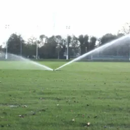 Automatyczny system nawadniania trawnika na boisku sportowym, z widocznymi zraszaczami rozpylającymi wodę w kształcie wachlarza, w tle drzewa i słupy oświetleniowe.