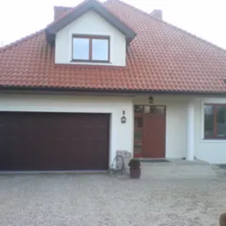 Dom jednorodzinny z brązową bramą garażową, drzwiami wejściowymi z szybami i oknami z brązowymi ramami pod czerwoną dachówką, oświetlony zewnętrzną lampą z numerem domu.