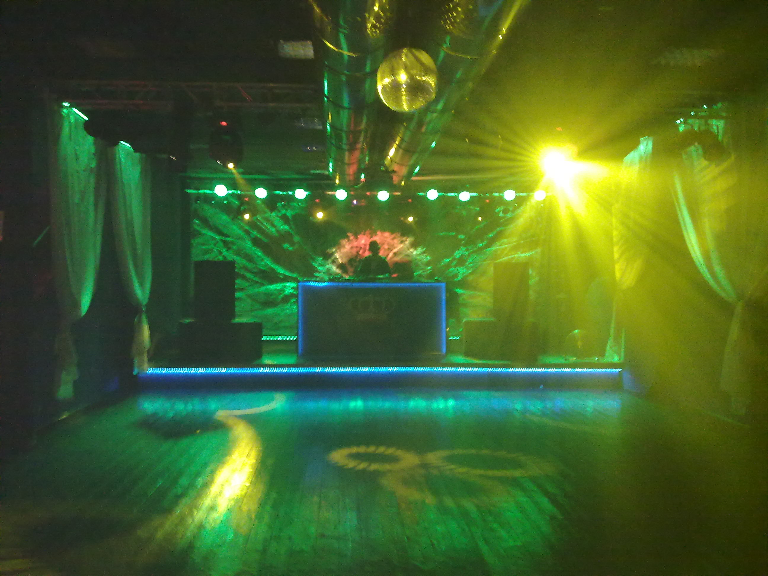 Scena oświetlona zielonymi i żółtymi światłami, z DJ-em za podświetlonym niebieskim światłem pulpitem, disco kula na suficie.