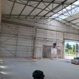 Dominik Marszałkowski Budownictwo Ogólne - Wnętrze hali w trakcie prac budowlanych: pracownik na rusztowaniu muruje otwór okienny, widoczne materiały budowlane, ściany z pustaków, konstrukcja dachu z metalowych belek, worek na śmieci...