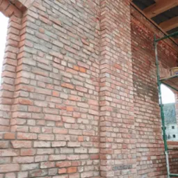 Murowana ściana z cegły klinkierowej z widocznym oknem w stanie surowym, fragment rusztowania i drewniana konstrukcja dachu.