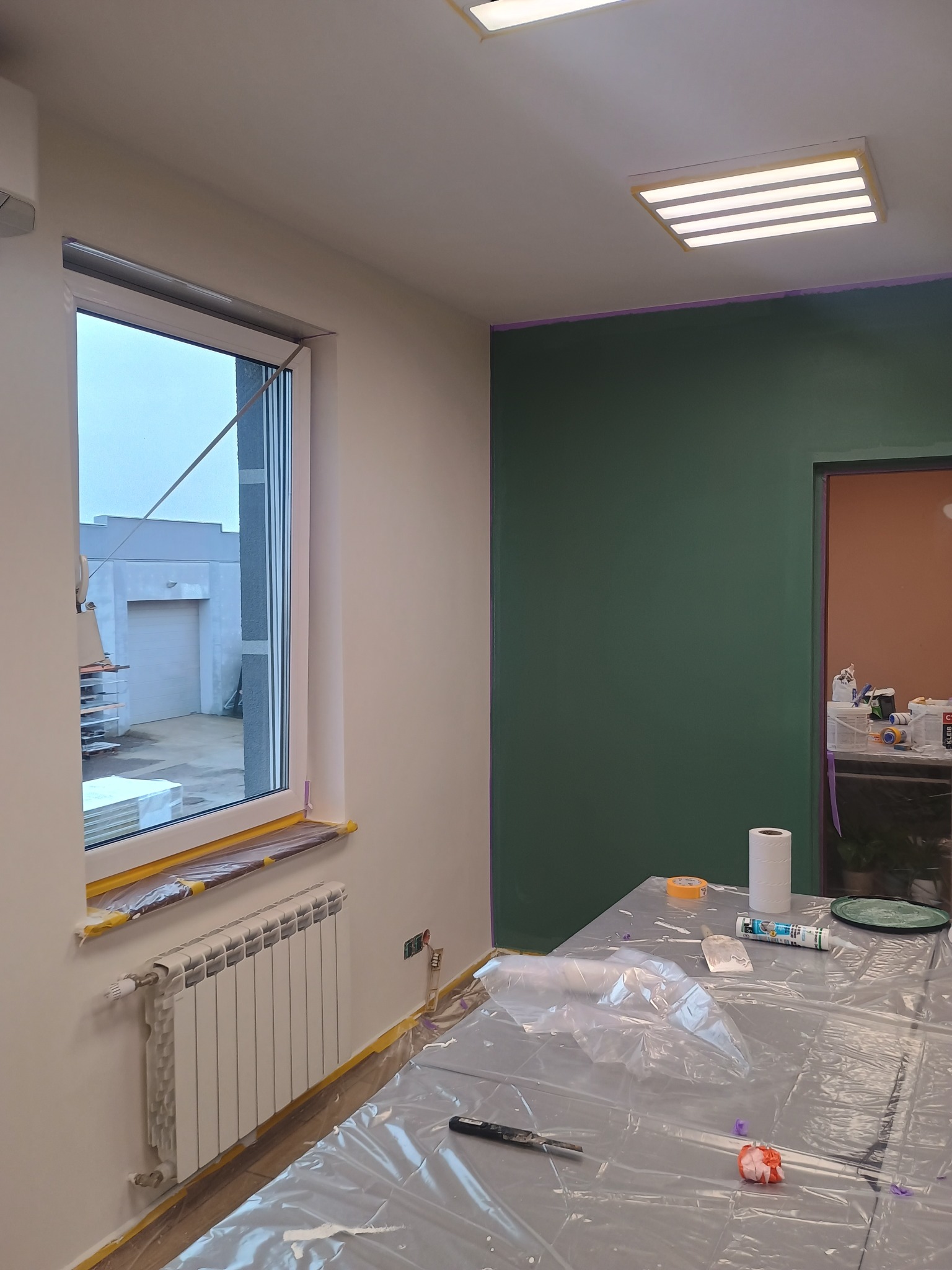 Pokój w trakcie malowania: zielona ściana, okno zabezpieczone folią, grzejnik, stół z narzędziami malarskimi przykryty folią ochronną.