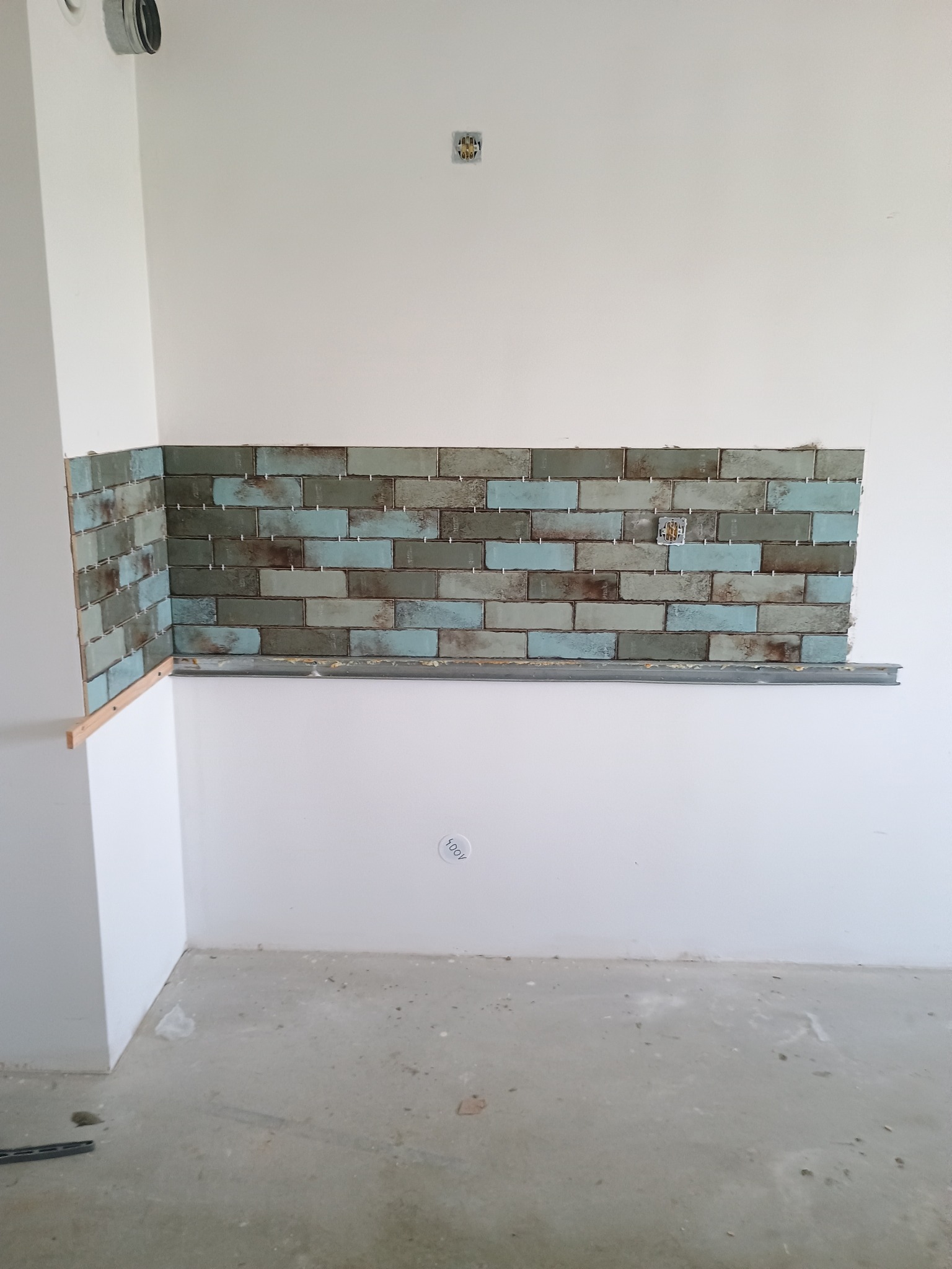 Fragment ściany z częściowo położoną, dekoracyjną glazurą w odcieniach brązu i błękitu, widoczne listwy montażowe i plastikowe krzyżyki dystansowe, surowy beton na podłodze.