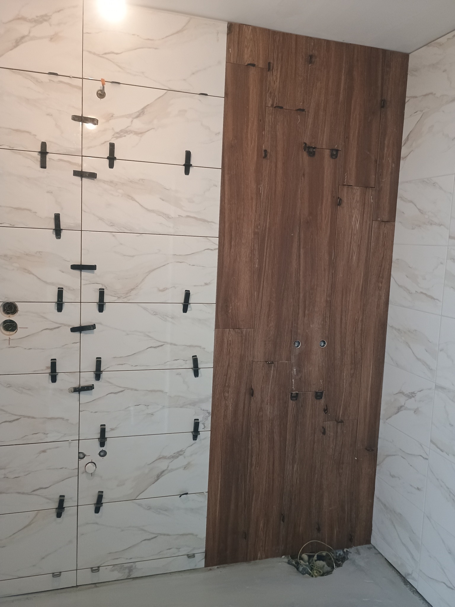 Ściana łazienki w trakcie układania płytek: białe płytki z marmurkowym wzorem po lewej stronie, drewnopodobne płytki po prawej, widoczne krzyżyki dystansowe i otwory na instalacje.