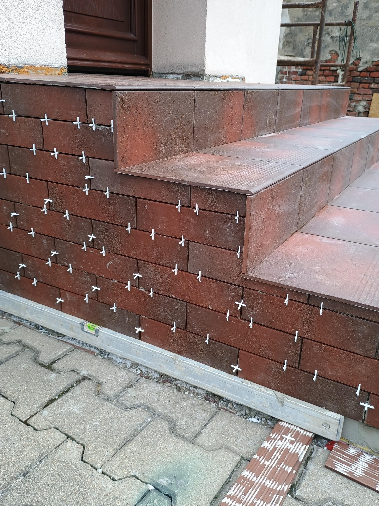 Wykończenie schodów wejściowych płytkami w odcieniach brązu, widoczne krzyżyki dystansowe i poziomica, na tle kostki brukowej i elewacji budynku.