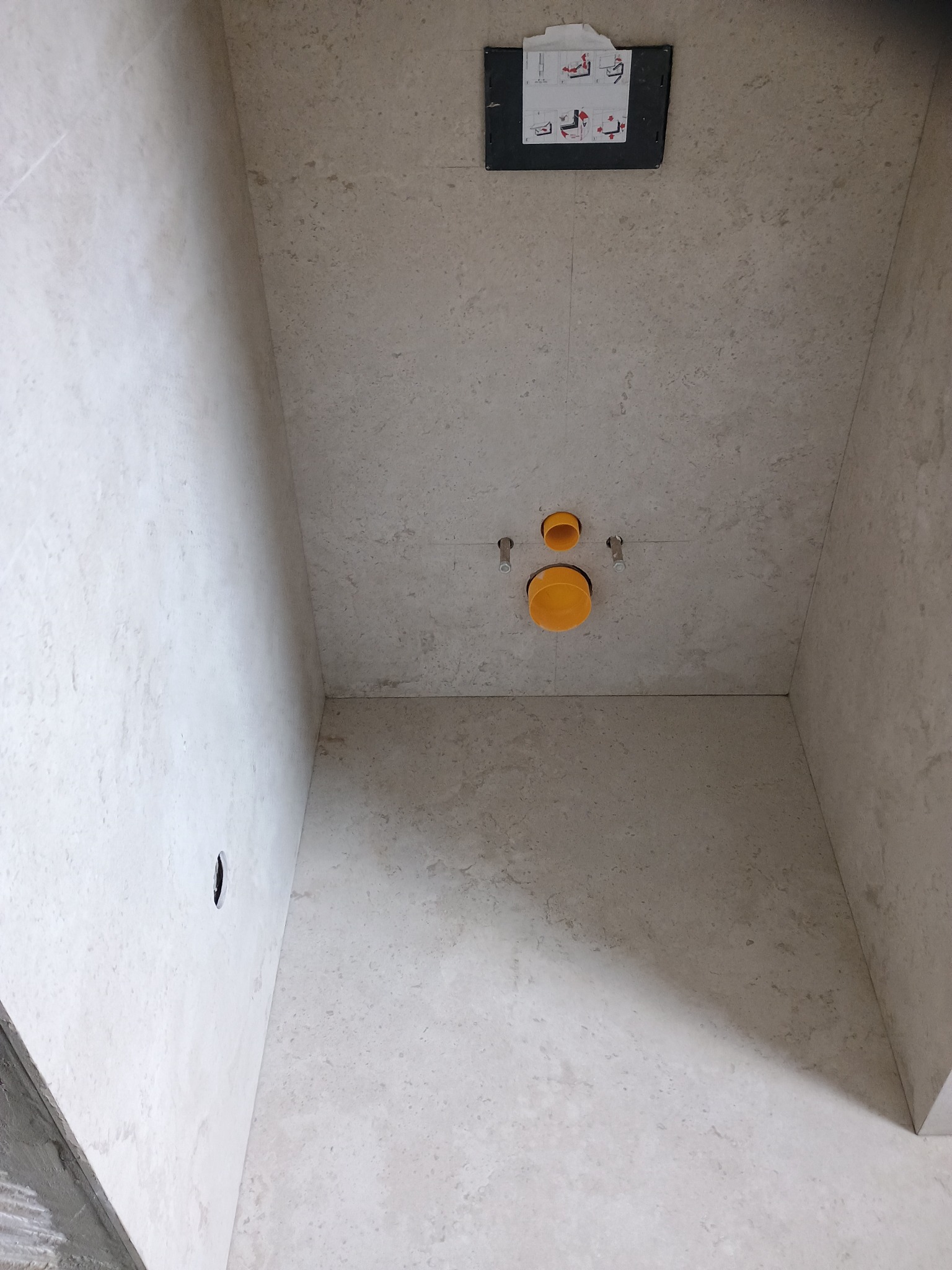 Wnęka w łazience wyłożona jasnymi płytkami imitującymi kamień, widoczne przygotowane przyłącza kanalizacyjne i wodne w kolorze pomarańczowym, zamontowana podtynkowa instalacja spłuczki WC...