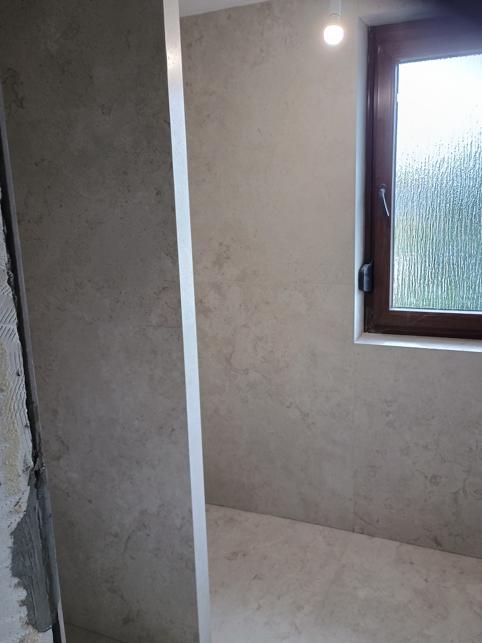 Ściana łazienki w trakcie wykańczania, widoczne płytki imitujące kamień, okno z szybą ornamentową i surowy fragment ściany z resztkami zaprawy.