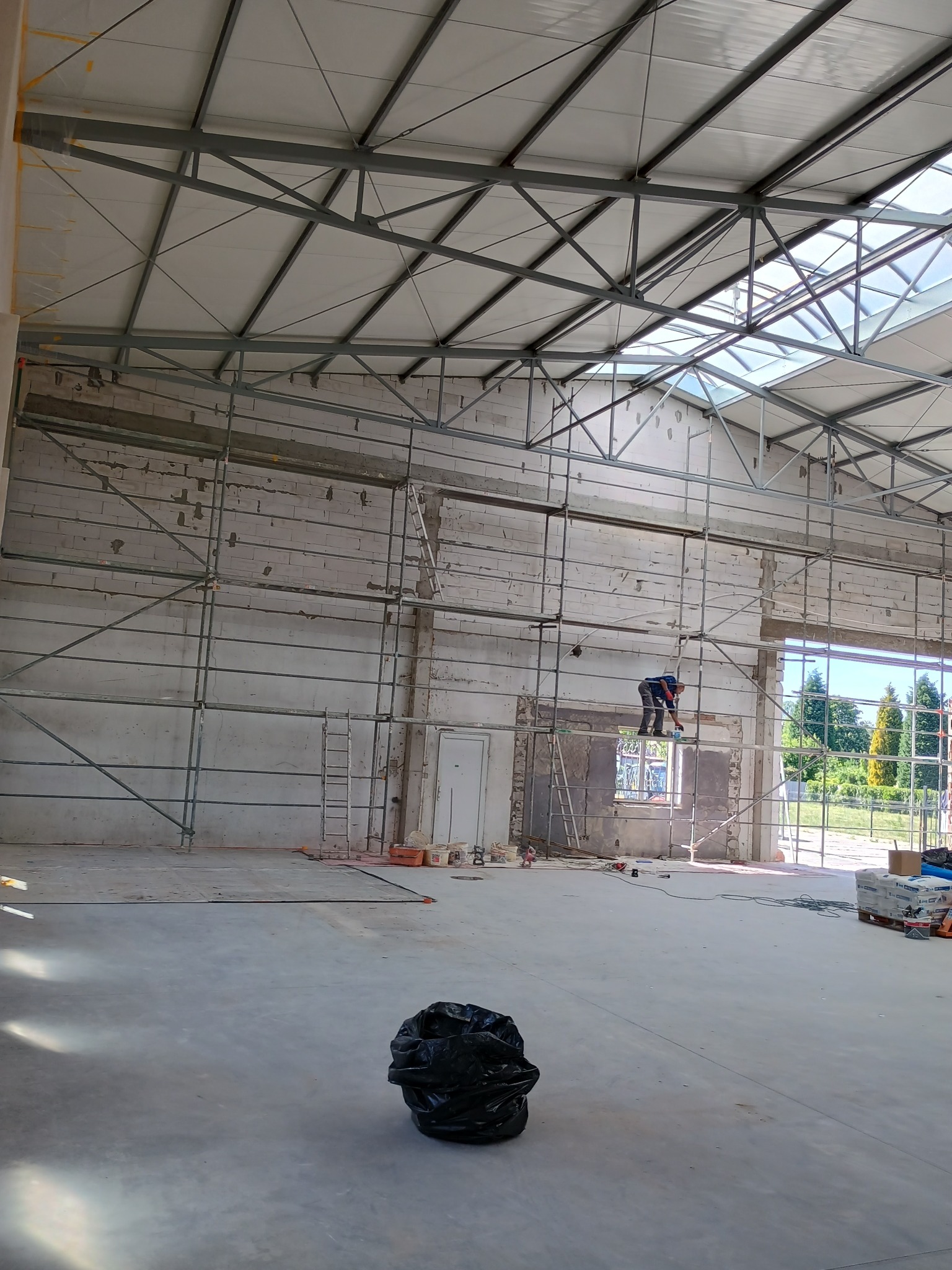 Wnętrze hali w trakcie prac budowlanych: pracownik na rusztowaniu muruje otwór okienny, widoczne materiały budowlane, ściany z pustaków, konstrukcja dachu z metalowych belek, worek na śmieci...