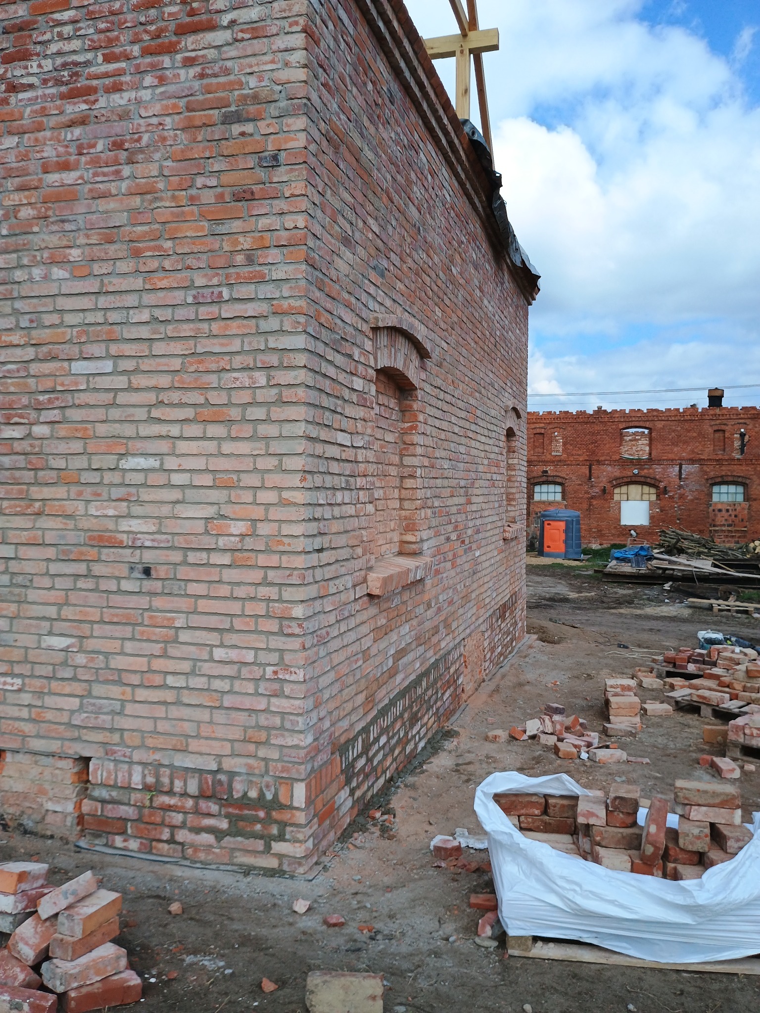 Wznoszony budynek z czerwonej cegły, widoczne fragmenty konstrukcji dachu i paleta z cegłami na placu budowy, w tle drugi budynek z cegły i przenośna toaleta.