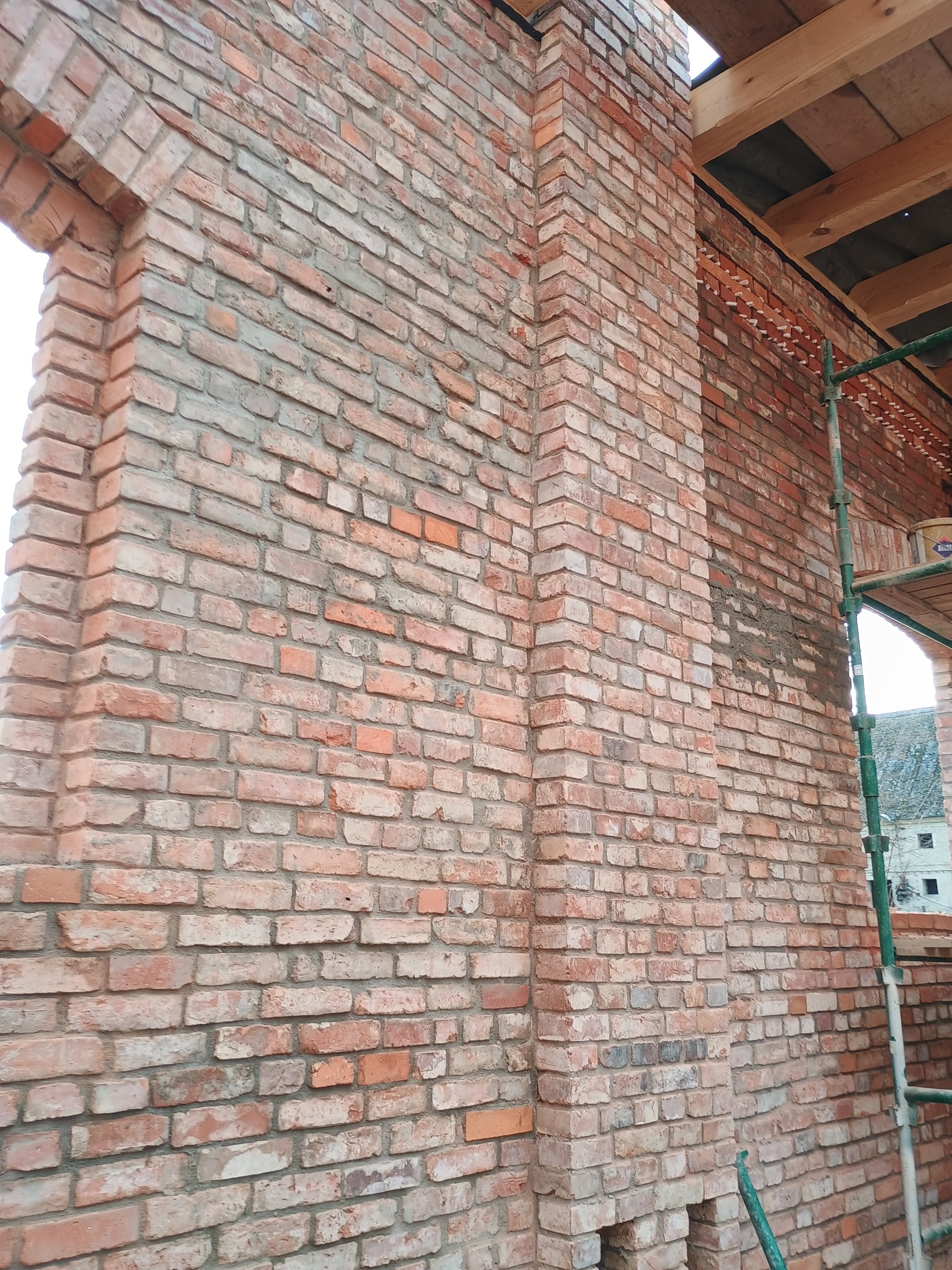 Murowana ściana z cegły klinkierowej z widocznym oknem w stanie surowym, fragment rusztowania i drewniana konstrukcja dachu.