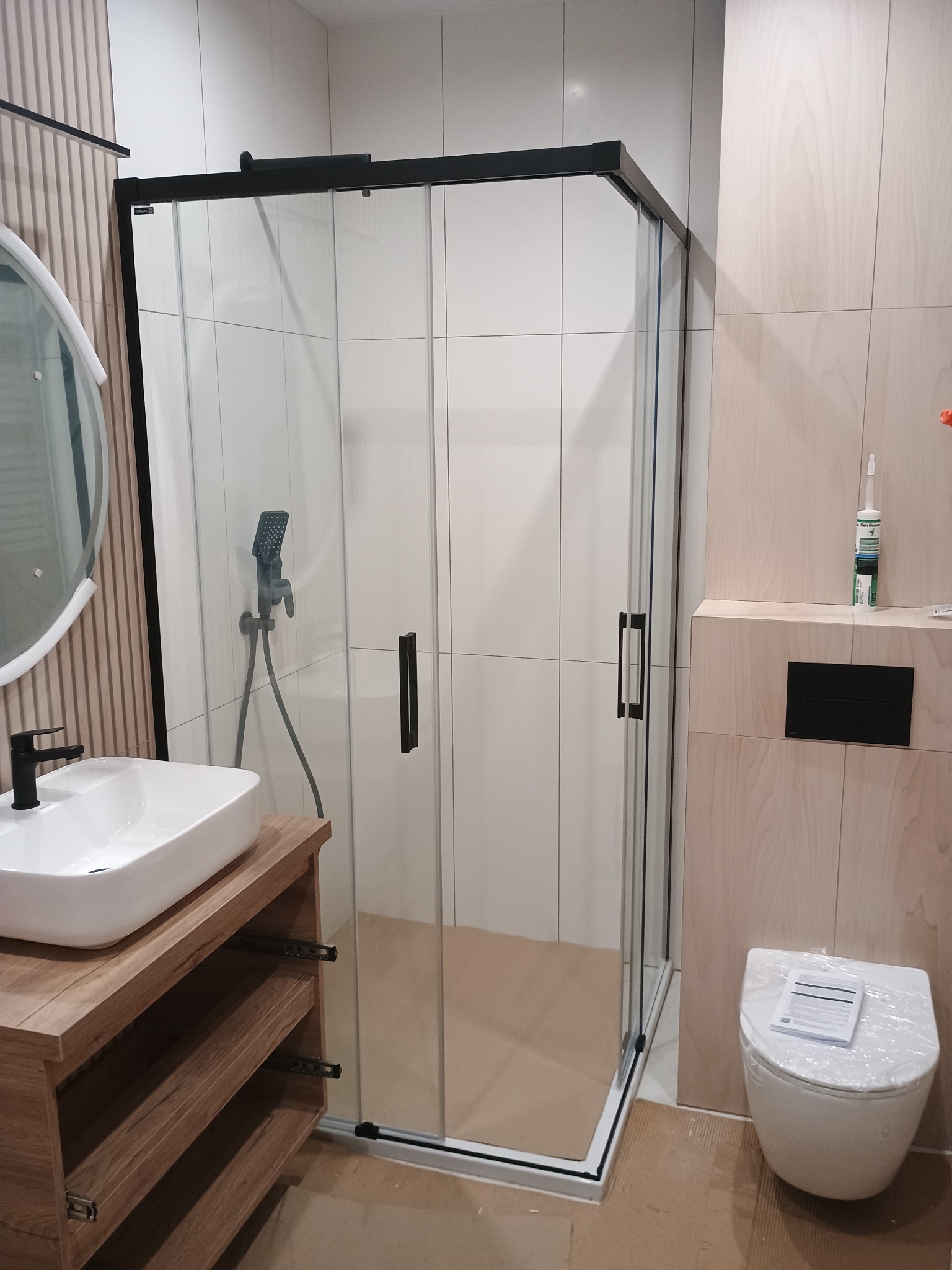 Nowoczesna łazienka z kabiną prysznicową typu walk-in z czarnymi profilami, umywalką nablatową na drewnianej szafce i wiszącą toaletą z czarnym przyciskiem.