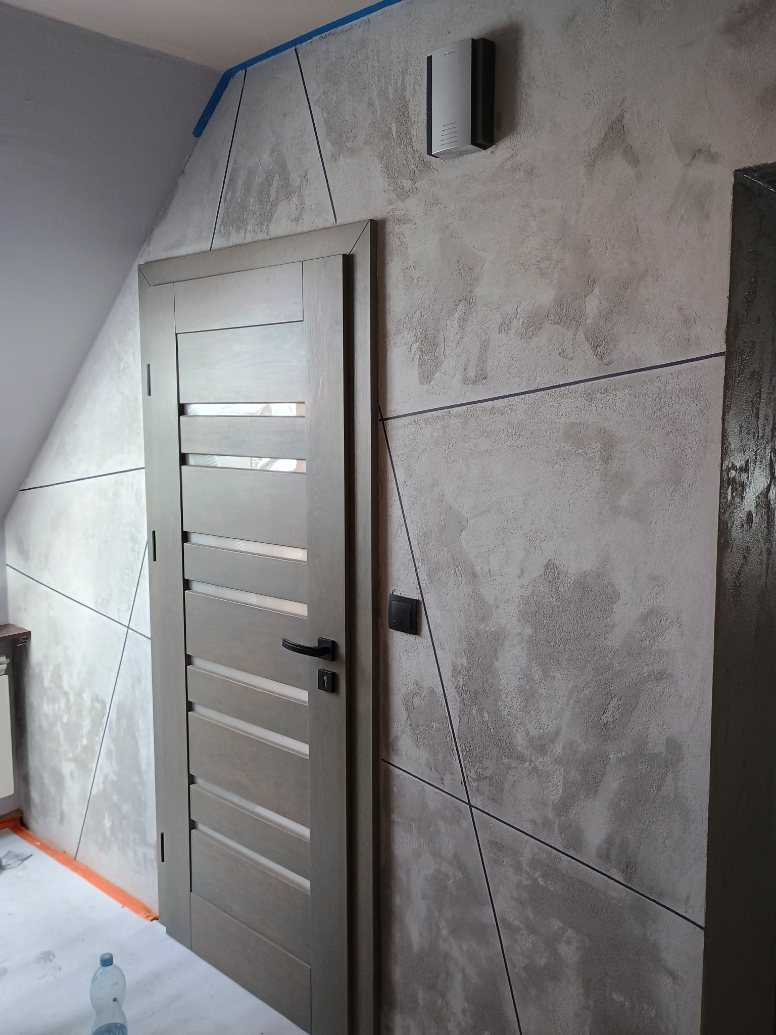 Wykończona ściana z efektem dekoracyjnym i geometrycznym wzorem, szara drewniana stolarka drzwiowa z czarną klamką, widoczny fragment poddasza.