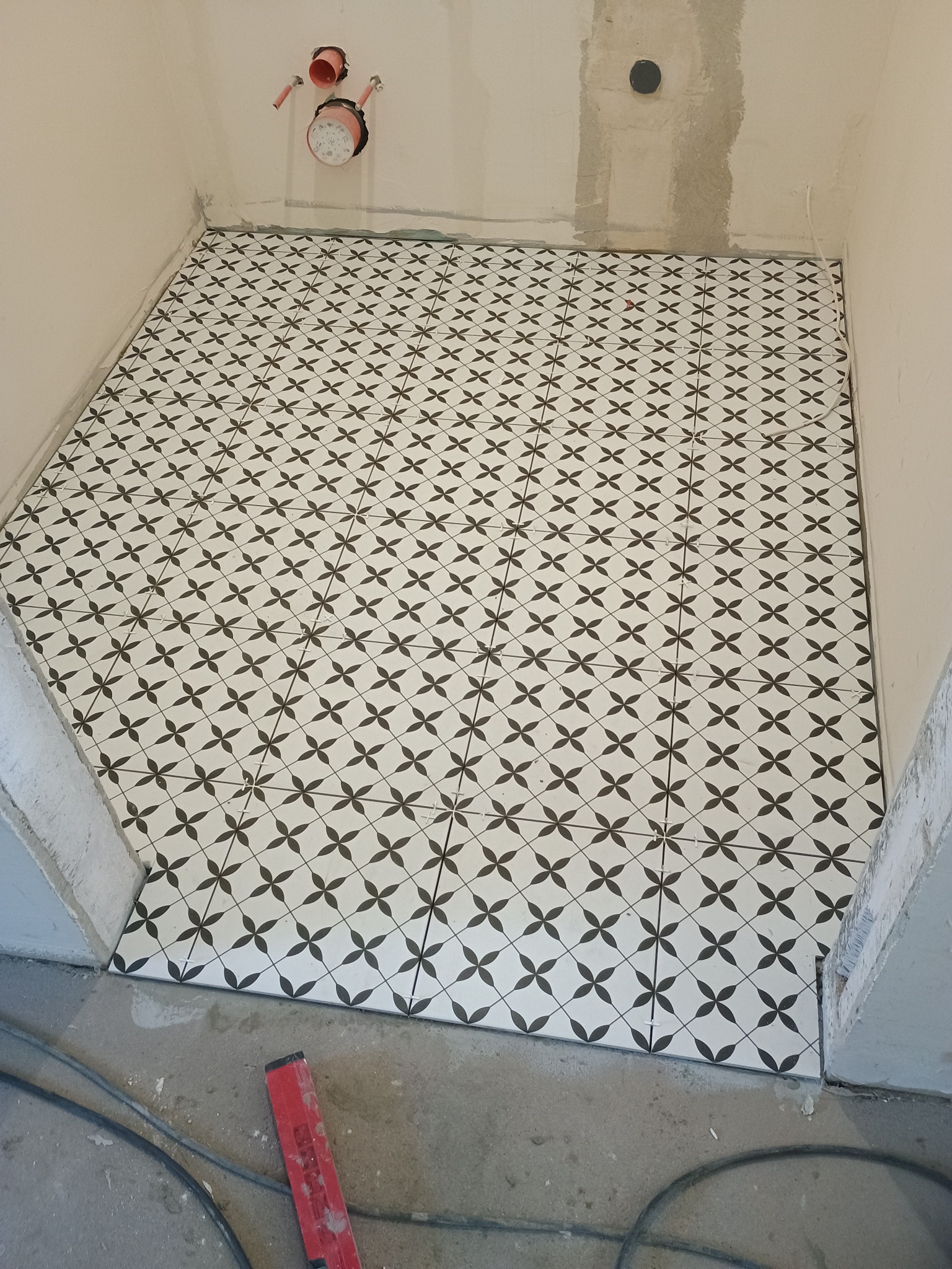 Ukończona podłoga w łazience z wzorzystych, czarno-białych płytek. Widoczne rury i poziomica. Świeżo położona podłoga w trakcie prac wykończeniowych.