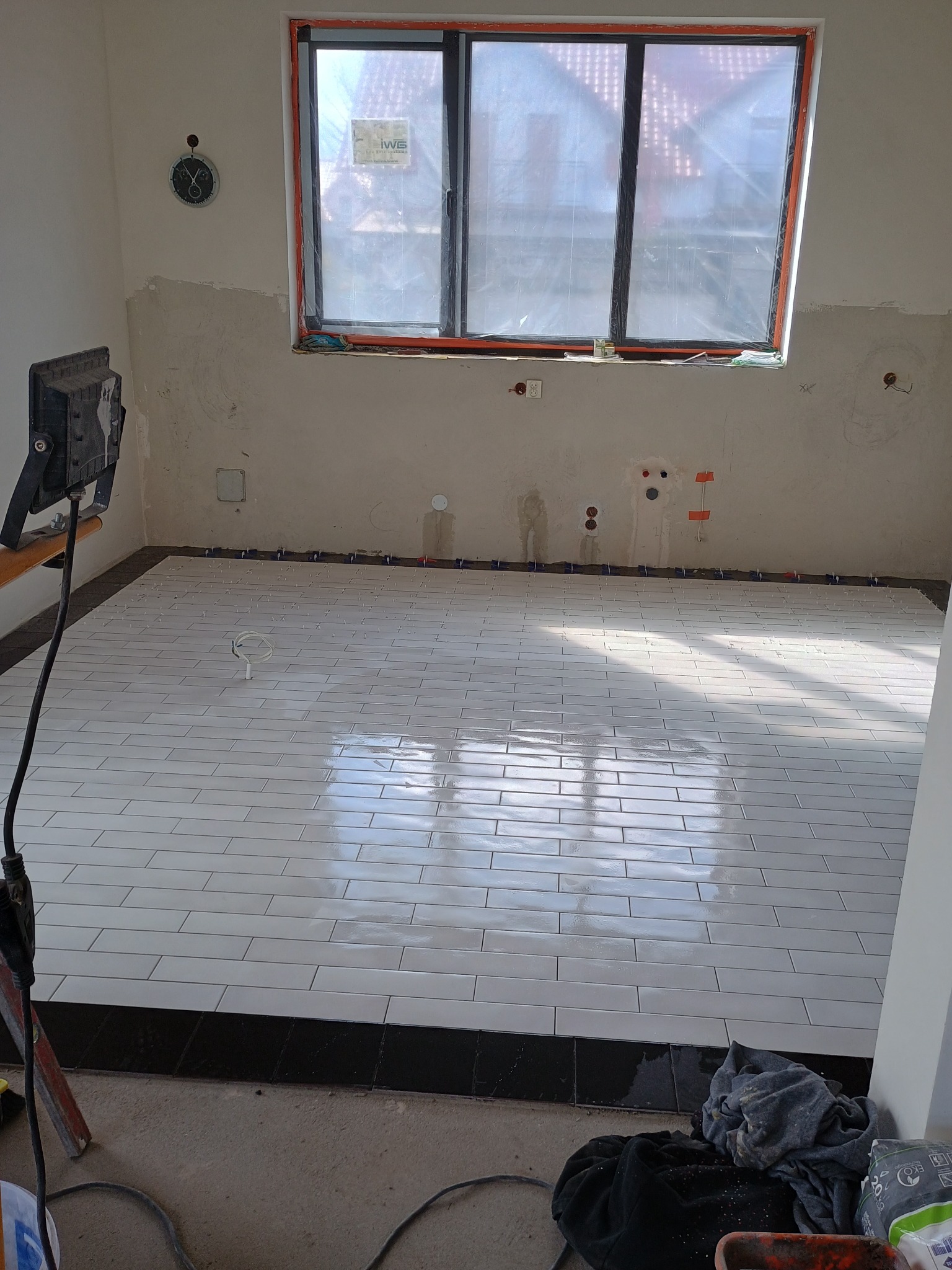 Ułożone białe, podłużne płytki na podłodze w trakcie remontu, z czarnym pasem płytek przy ścianie. Widoczne krzyżyki dystansowe, ściany przygotowane do dalszych prac.