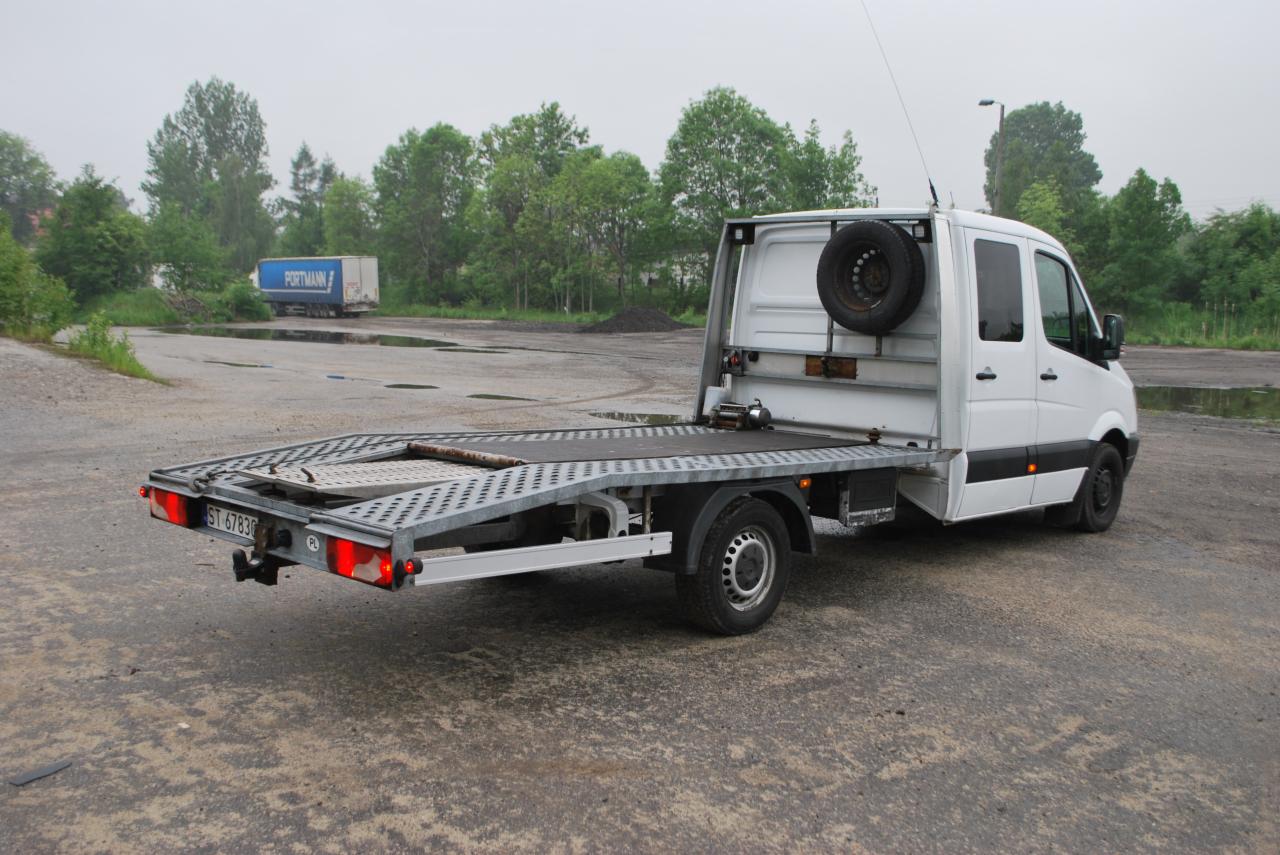 Biała laweta z platformą załadunkową na szarym placu, z zapasowym kołem na tylnej ścianie kabiny. W tle widoczna naczepa z napisem PORTMANN.