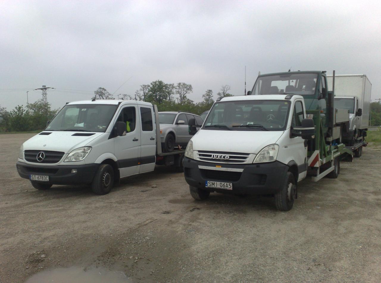 Dwie białe lawety: Mercedes Sprinter i Iveco, zaparkowane na szutrowym placu. Na jednej z nich przewożony jest srebrny samochód osobowy i kabina ciężarówki.