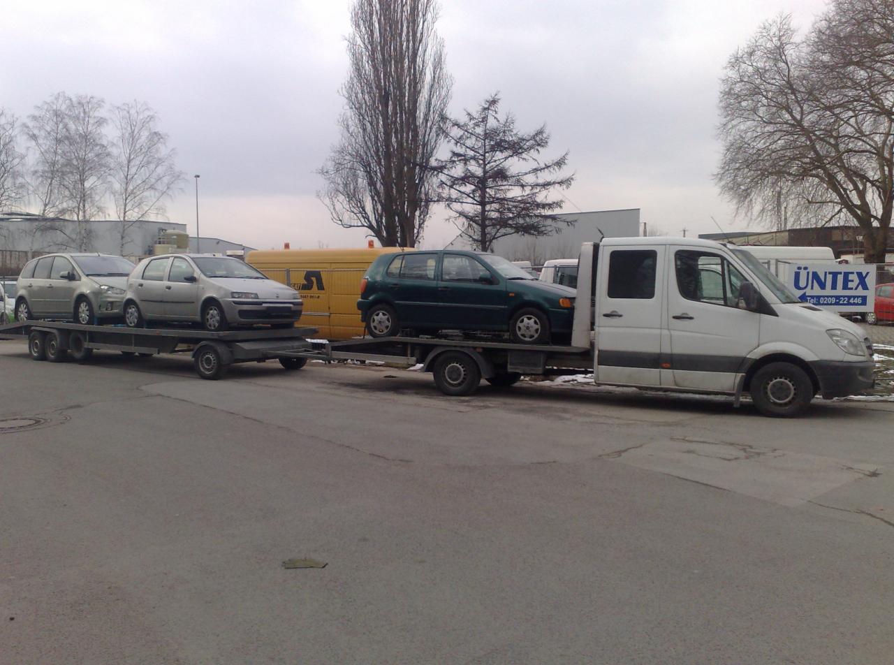 Biała laweta z trzema samochodami osobowymi na przyczepie, zaparkowana na szarej drodze w pochmurny dzień. Widoczny żółty samochód serwisowy w tle.