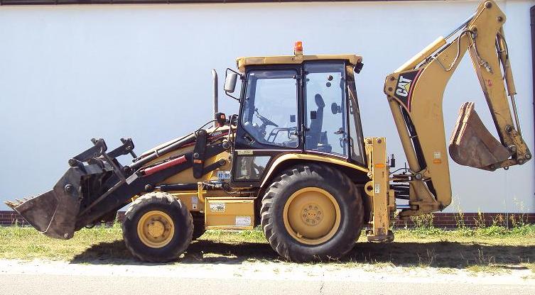 Żółto-czarna koparko-ładowarka CAT, zaparkowana na trawie przed białym budynkiem, widok z boku, z łyżką z przodu i z tyłu.