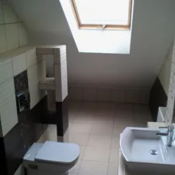 Wykończona łazienka na poddaszu ze skośnym sufitem i oknem dachowym, jasne płytki z ciemnobrązowymi akcentami, podwieszana toaleta z zabudową i umywalka z lustrem.