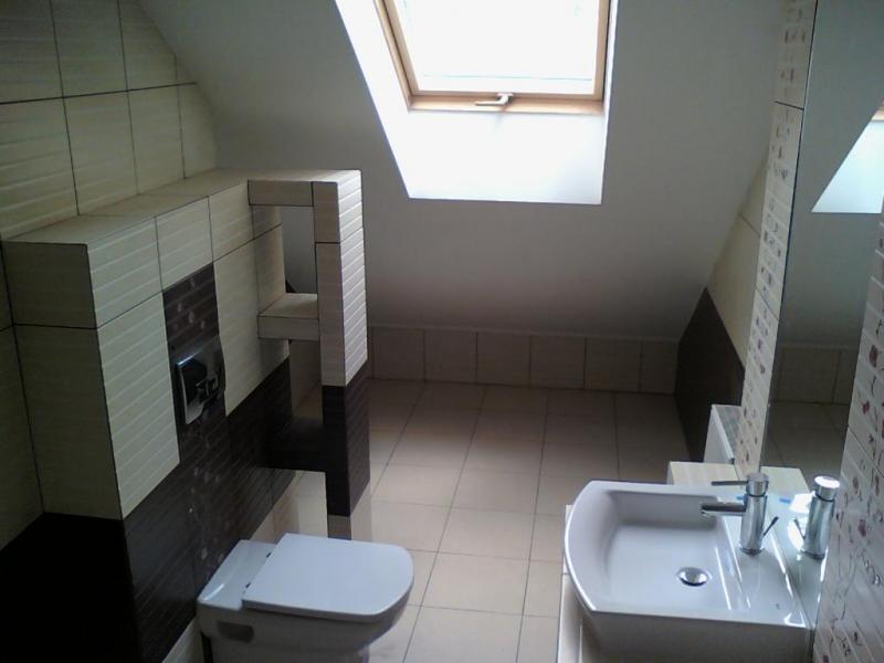 Wykończona łazienka na poddaszu ze skośnym sufitem i oknem dachowym, jasne płytki z ciemnobrązowymi akcentami, podwieszana toaleta z zabudową i umywalka z lustrem.