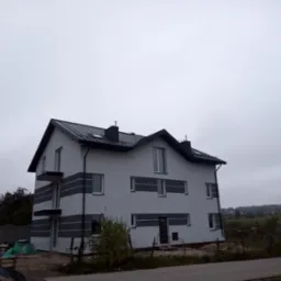 Dwukondygnacyjny dom z szarą elewacją, czarnym dachem i balkonami, widoczny z perspektywy ulicy w pochmurny dzień, z elementami placu budowy w tle.