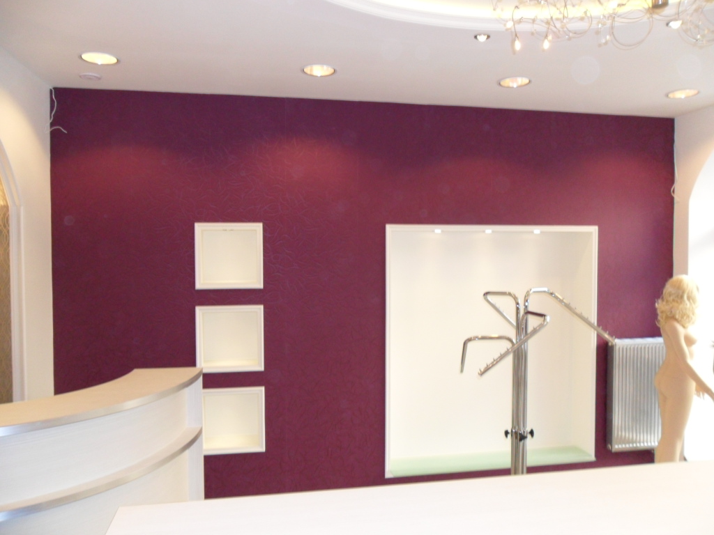 Wnętrze sklepu odzieżowego z bordową ścianą, wnękami z oświetleniem, manekinem i wieszakiem na ubrania, oświetlone halogenami i ozdobnym żyrandolem.